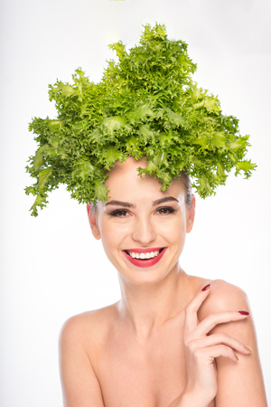 Happy young woman is standing with lettuce on her head. She is looking at camera with smiling. Isolatedの写真素材