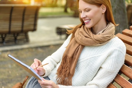 Joyful female artist is creating picture on nature. She is sitting on bench and smiling