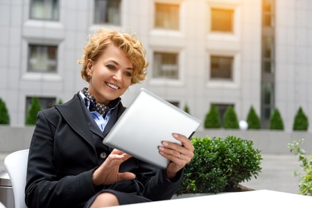 Elegant young woman is using tablet and smiling. She is sitting at table in cafe outdoorsの写真素材