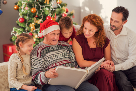 This is our family album. Parents and children are looking at photos with interest. They are sitting on sofa near Christmas tree and smilingの写真素材