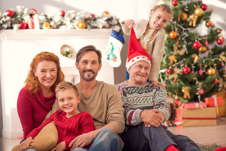 We are happy together. Joyful married couple is sitting on sofa and embracing their son. Girl is touching Christmas hat of grandfather and smilingの写真素材