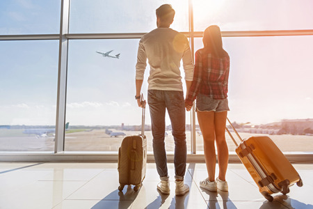 Inspired young loving couple is looking at flying plain in sky. They are standing near window at airport and holding hands Inspired young loving couple is looking at flying plain in sky. They are standing near window at airport and holding hands