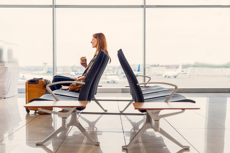 Packed and prepared for take-off. Beautiful smiling woman enjoying cup of coffee while waiting for landing at airportの写真素材