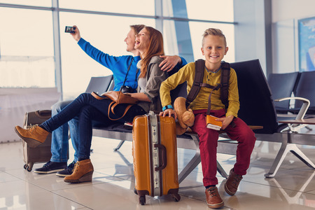 Time with family is time well spent. Little smiling boy sitting at airport and holding tickets in his hand with his parents making selfie in backgroundの写真素材