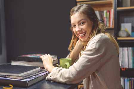 Books are my passion. Happy beautiful girl sitting at desk with lots of books and enjoying coffeeの写真素材