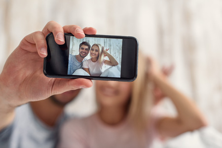 Joyful loving couple is making selfie in bed. They are sitting and smiling. Focus on smartphone