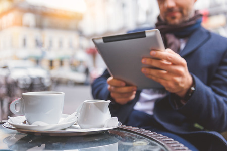 Relaxed man is sitting in cafe on street and holding tablet. Focus on cup of coffee and milk jar on tableの写真素材
