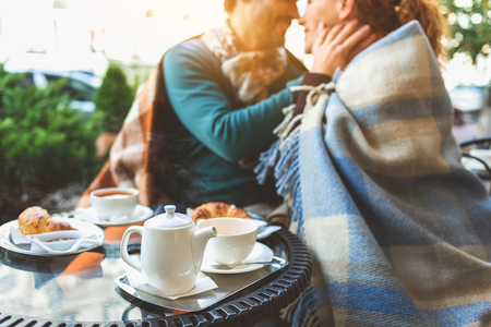 Loving couple is sitting and hugging. They are looking at each other with love and smiling. Focus on tea and croissant on tableの写真素材