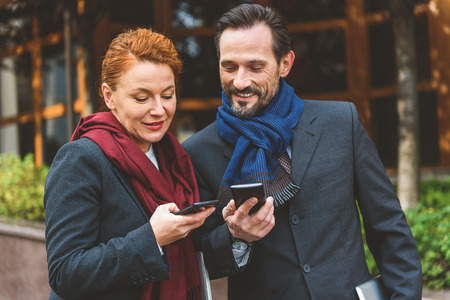 Middle-aged man and woman are exchanging phone numbers. They are standing on street and laughingの写真素材