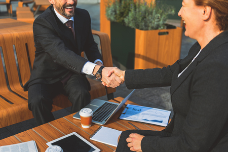 Beginning partnership. Happy businessman and businesswoman are shaking hands and smiling. They are sitting in cafe outdoorsの写真素材