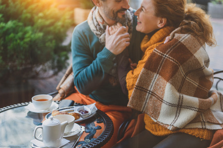 Happy married couple is warming up by their love in autumn. They are sitting at table and hugging. Lovers are smilingの写真素材