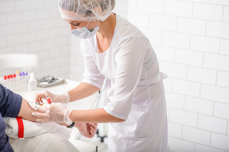 Experienced female doctor is undergoing blood test for man in clinic. She is standing and taking needle from his vein accuratelyの写真素材
