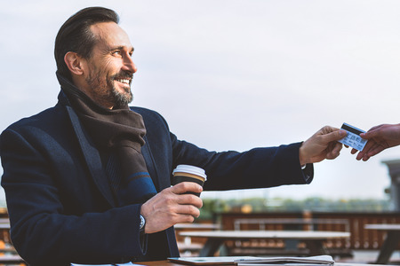 Happy middle-aged man is playing for coffee by credit card in cafe. He is sitting outdoors and smilingの写真素材