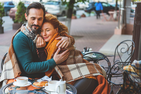 Joyful man and woman are sitting at table in cafe outdoors and embracing with love. They are smilingの写真素材