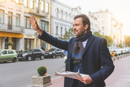 Successful businessman is calling for taxi in city. He is raising arm and smiling. Worker is standing and holding newspaperの写真素材