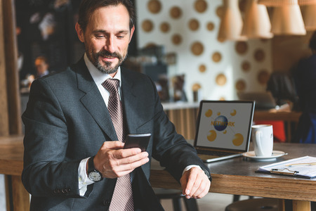 Happy man is messaging on mobile phone and smiling. He is sitting in restaurant near laptopの写真素材