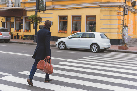 Businessman is crossing the street in a hurry. He is carrying bag. Focus on his backの写真素材