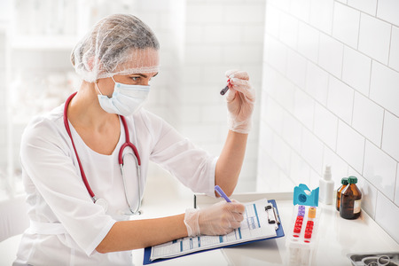Professional female scientist is looking at blood test tube with seriousness. She is sitting at desk and notingの写真素材