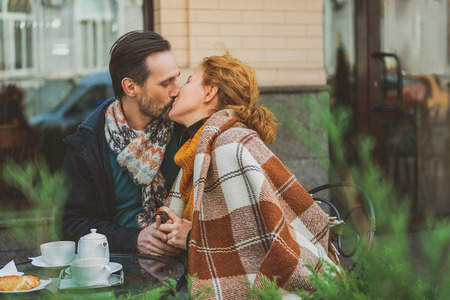 Happy couple is kissing with love. They are sitting at table in cafeteria outdoors and holding handsの写真素材