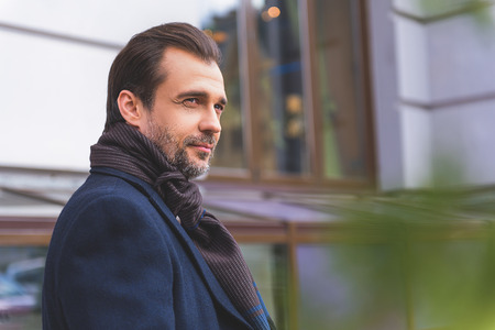 Thoughtful man is standing outdoors in town. He is wearing coat and scarfの写真素材