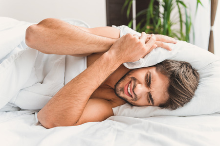 Man is lying on left side, screwing his eyes. He is covering ears by pillow beacause of noiseの写真素材