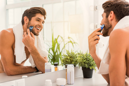 Morning begins with shaving. Joyful young man is looking at mirror and smiling. He is standing and carrying white towelの写真素材