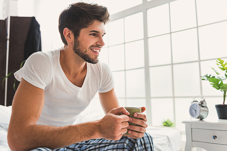 Happy young man is enjoying coffee in morning. He is sitting on bed and smilingの写真素材