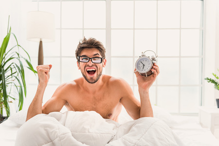 I oversleep. Young man is sitting on bed and holding alarm-clock. He is screaming with shockの写真素材