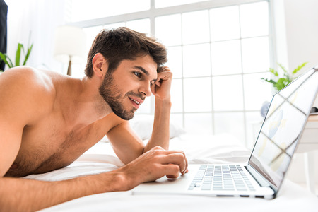 Carefree young man is communicating in internet with aspiration. He is lying on bed and laughingの写真素材