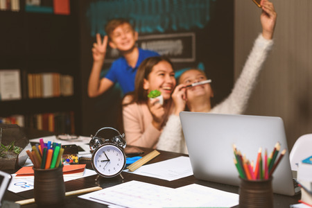 Focus on working table with computer, clock and equipment for learning. Children are making selfie...