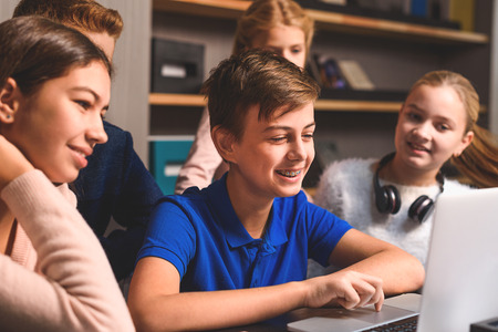 Vigorous boy is using laptop and other children are watching his with smile