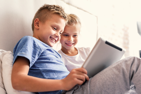 Growing up with technology. Two little smiling kids sitting on bed at home and playing games on their technology tabletの写真素材