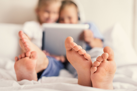 High tech story time. Close up little smiling boy and girl sitting on bed with digital tablet, focus on feetの写真素材