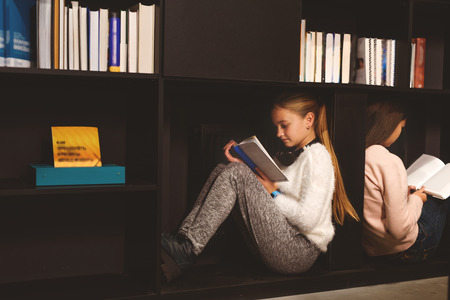Two fascinated girls are sitting on shelves with open volumesの写真素材