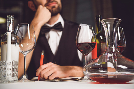 Thoughtful man is sitting at table and holding pen. Focus on carafe and glasses with scarlet drinkの写真素材