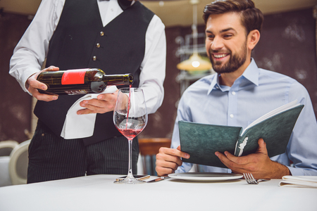 Accurate waiter pouring scarlet nectar into glass. Smiling man is sitting at table and expecting his wineの写真素材
