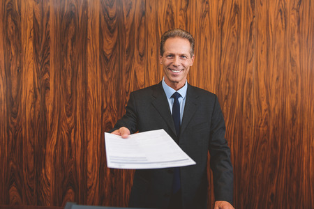 You must fill out form. Smiling male mature receptionist standing behind counter and giving documents for signatureの写真素材