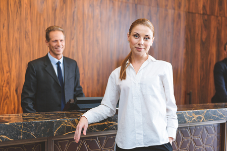 Waiting for porter. Smiling young woman standing near counter desk and looking at camera with hotel manager on backgroundの写真素材