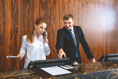 Be attentive. Female receptionist talking on phone with customer while male desk clerk pointing fingert at computer screenの写真素材