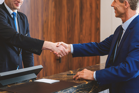 Welcome to apartments. Smiling male receptionist standing behind counter and giving handshake to senior guestの写真素材