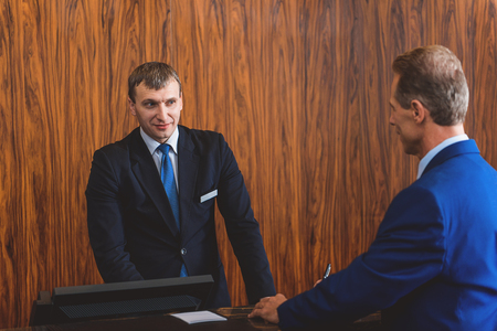Can I help you. Adult businessman in suit talking to smiling hotel worker while standing in front of hotel reception deskの写真素材