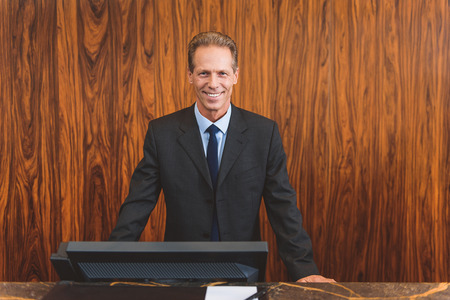 With the best service. Friendly mature male receptionist standing behind counter in hotel lobby with welcoming smileの写真素材