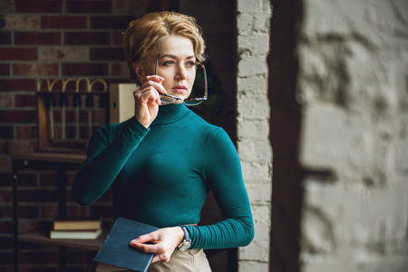 Thoughtful woman is standing in room. She looking at side, holding notebookの写真素材