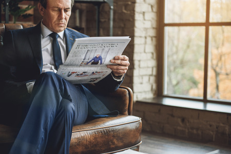 Concentrated mature man is sitting on sofa in official suit. He looking through daily newspaperの写真素材