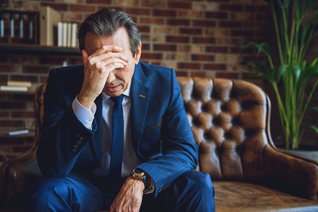 Worried mature businessman is sitting on couch. He touching his forehead with hand and closing eyesの写真素材
