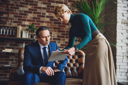 Serious matured businessman is sitting on couch while psychologist standing beside him and...