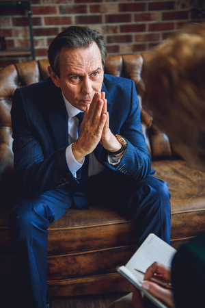 Tired senior businessman is sitting on couch. He showing request sign.の写真素材