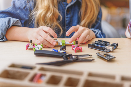 Small inventor is sitting at board. She holding spare parts of some mechanism