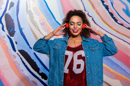 Happy african female with blue eyeshadows listening music in earphones near pictureの写真素材