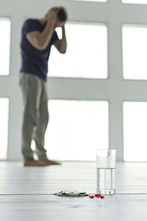 Focus on small scarlet capsules near glass with beverage. Upset man standing nearby window. Drug addiction conceptの写真素材